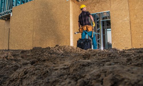 Construction worker wearing a yellow hard hat and tool belt, standing inside a partially built structure with exposed wooden and metal framework.
