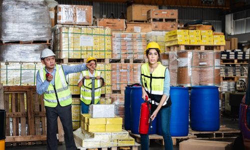 Warehouse workers in safety gear manage logistics, with a supervisor directing while a female worker holds a fire extinguisher among stacked goods and barrels.